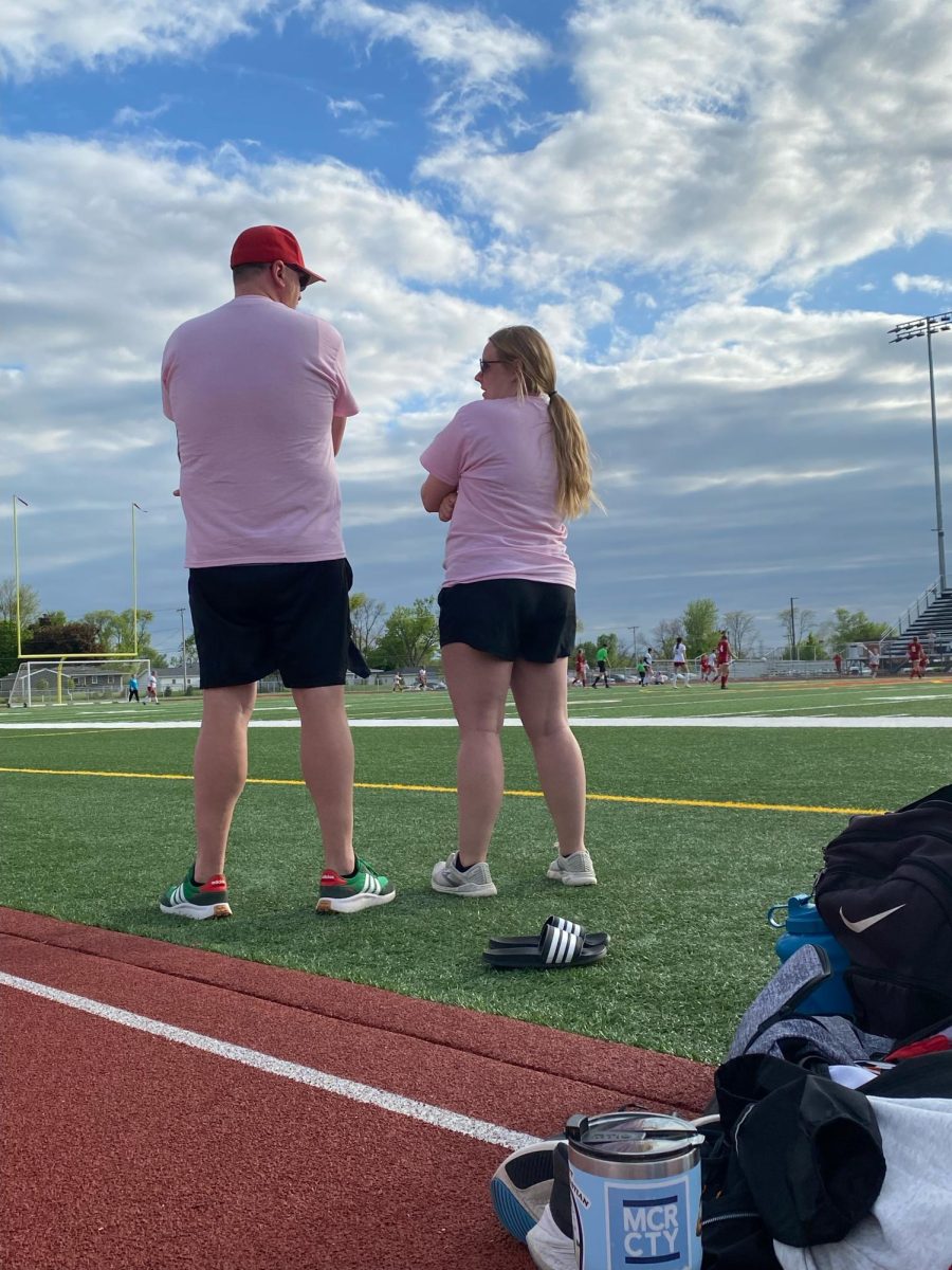 Coach Trilk and Coach Vega stand on the sidelines overlooking her players