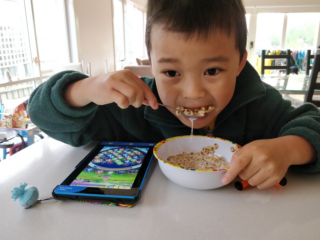 A kid eats his cereal while playing games on his iPad.